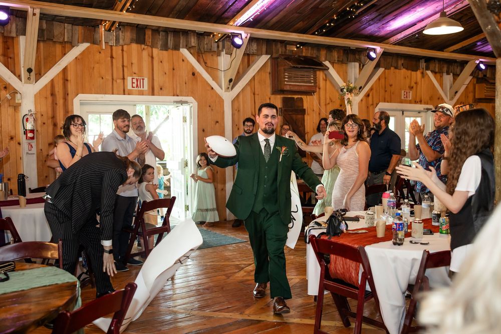 Groom in green suit makes grand entrance at rustic barn wedding reception with guests cheering around him.
