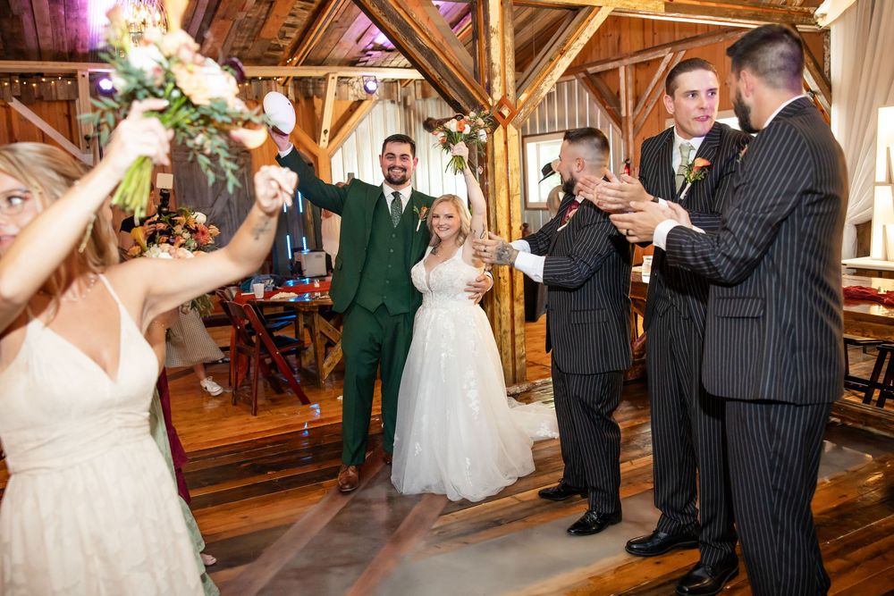 Newlywed couple in green suit and white gown celebrate wedding reception entrance in rustic barn venue with cheering guests.