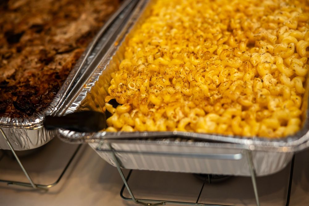 Baked macaroni and cheese in a large aluminum catering tray, golden and cheesy, served alongside a meat dish.