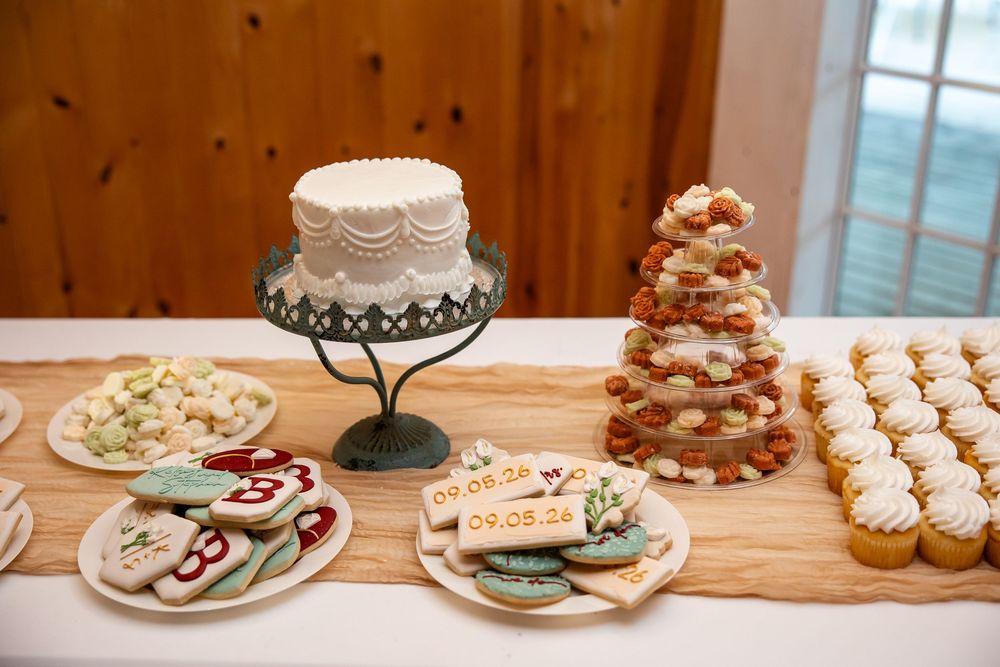Elegant dessert table with white wedding cake, decorated cookies, donut tower, and cupcakes on a rustic wooden surface.