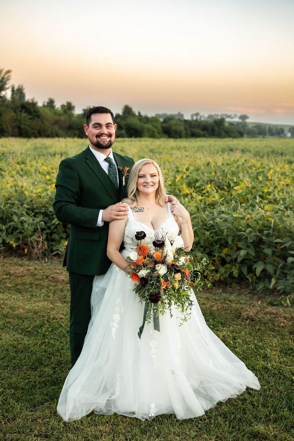 Bride and groom posing at sunset in a lush green field, bride holding a colorful bouquet, groom in a dark green suit.
