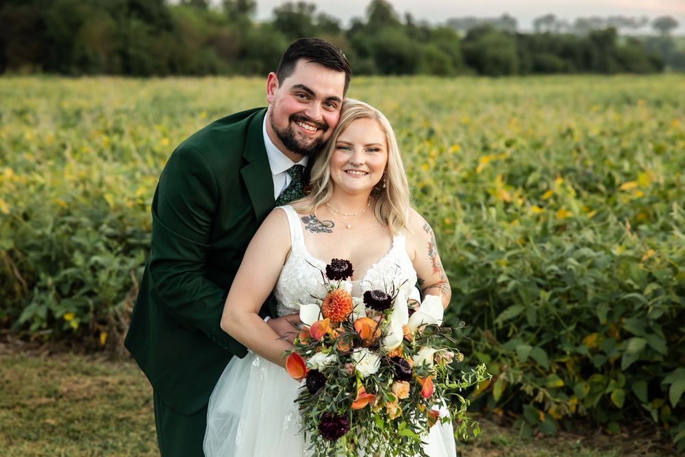 Smiling bride holding colorful bouquet with groom in green suit posing in front of lush green farm field.