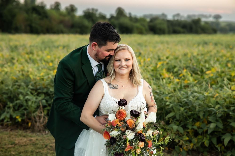 Bride holding colorful bouquet with groom in green suit embracing her in front of sunflower field at outdoor wedding.