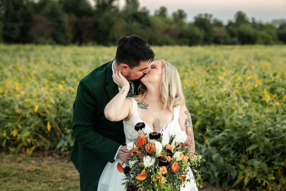 A bride and groom share a romantic kiss in a sunflower field, she holds a vibrant autumn bouquet.
