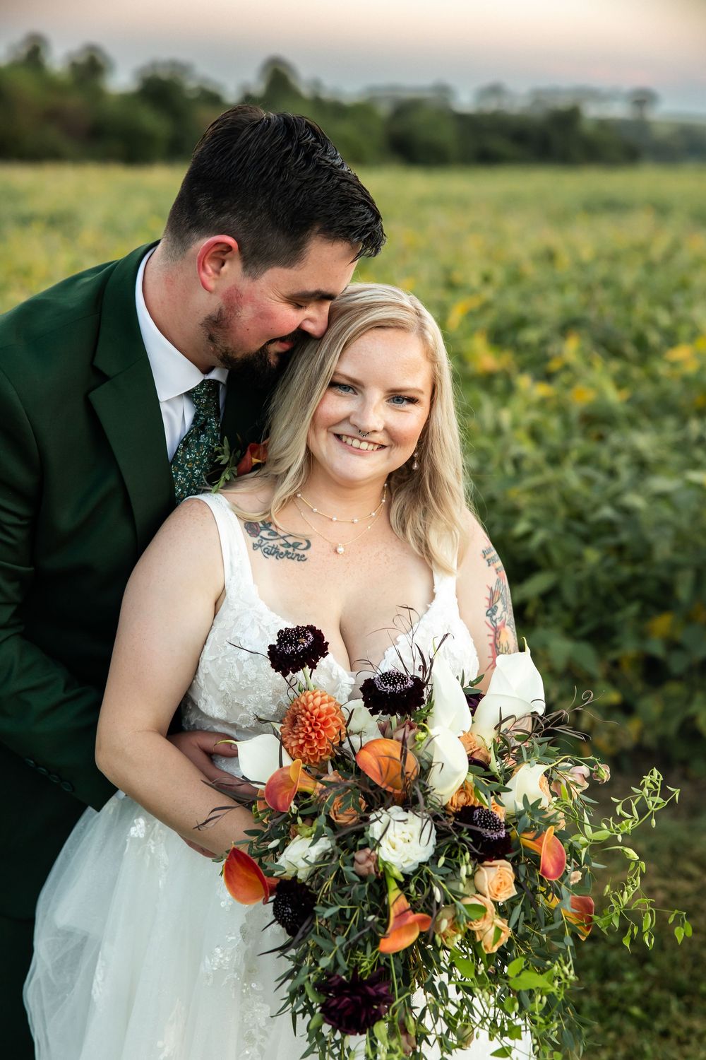 Bride in lace gown holds colorful bouquet with groom in green suit, posing in a sunflower field at sunset.