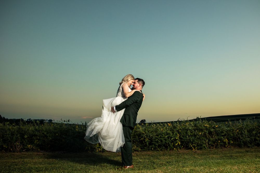 Groom lifts bride in white gown at sunset, sharing a romantic kiss outdoors near a vineyard.