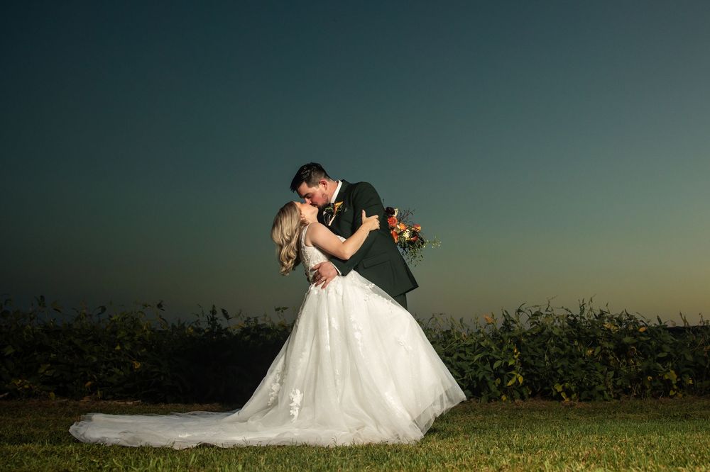 Bride and groom share a romantic dip kiss at sunset in an outdoor wedding portrait with a glowing twilight sky.