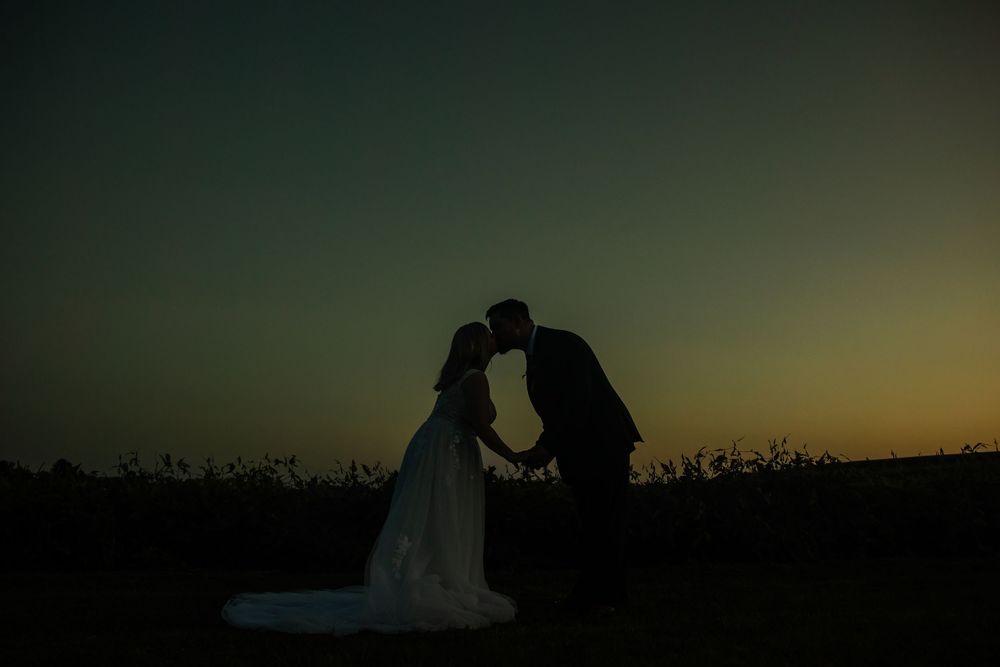 Silhouette of a bride and groom sharing a romantic kiss at sunset in an outdoor field.