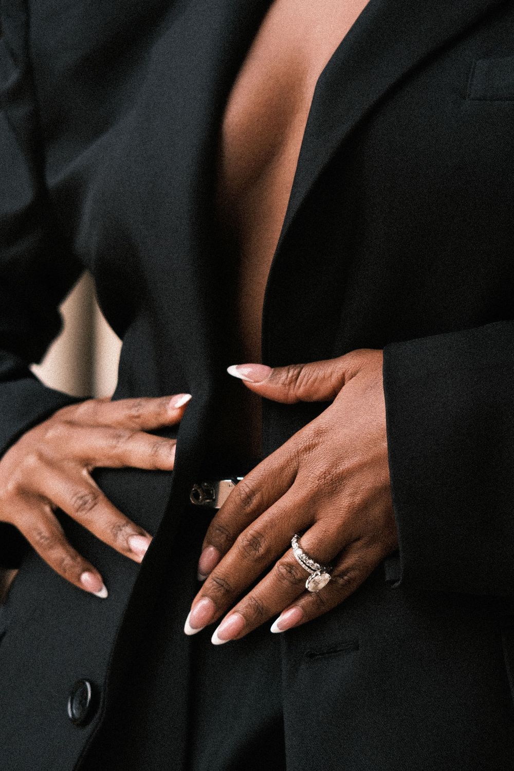Close-up of manicured hands with pink nails and a silver ring resting on a black tailored blazer worn open.