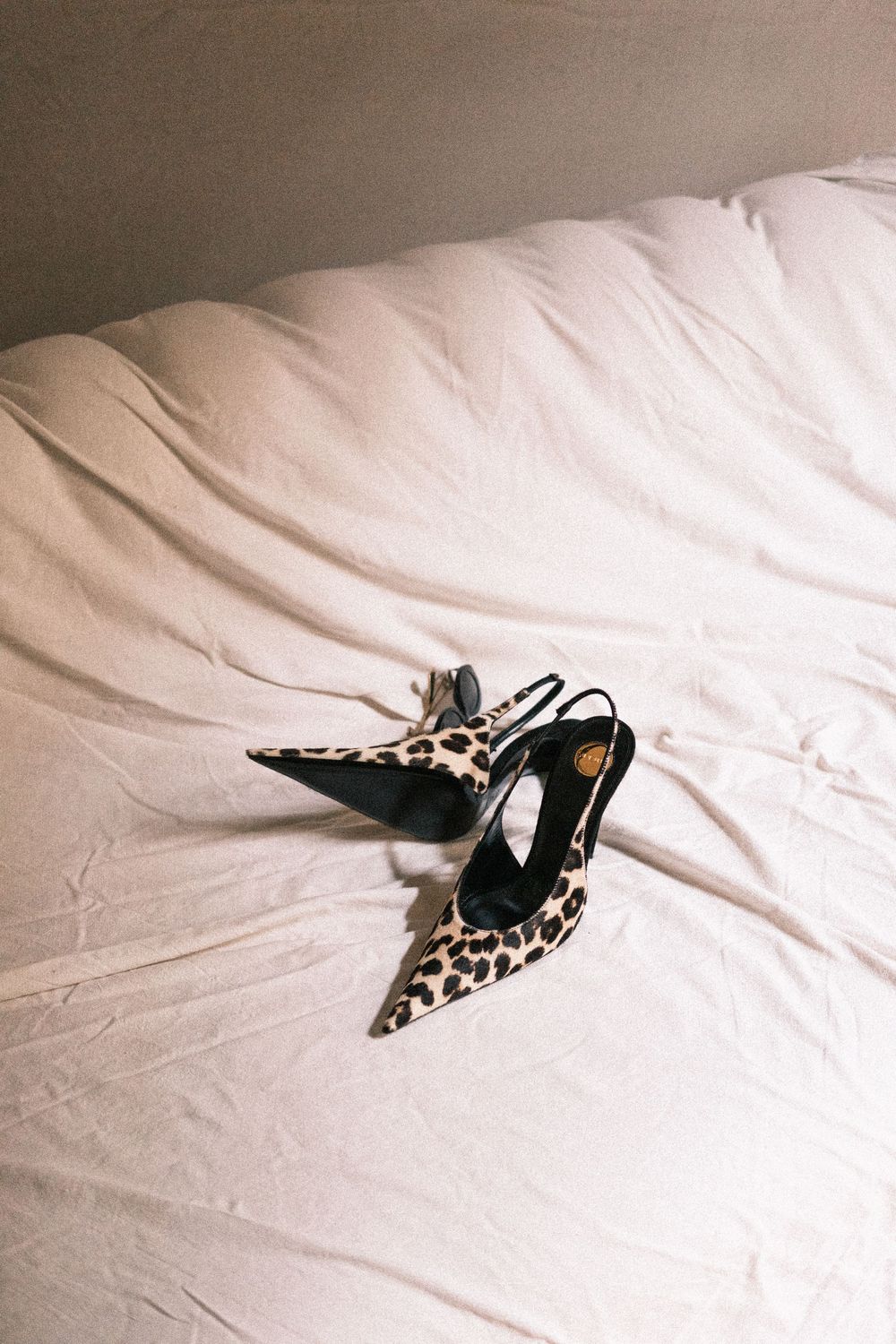 Pair of leopard print slingback pointed-toe stiletto heels with black accents resting on rumpled cream bed linens.