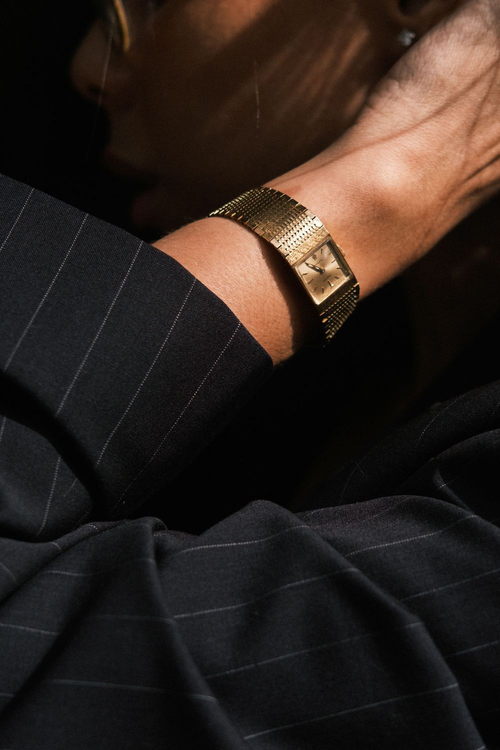 Close-up of a gold mesh bracelet watch on a wrist, paired with a dark pinstripe suit jacket.