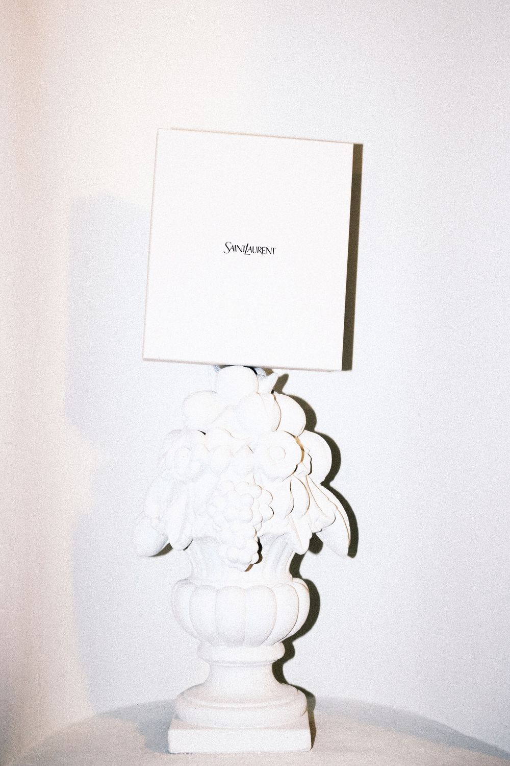 White sculptural vase with floral details holding a Saint Laurent branded white box on a marble surface.