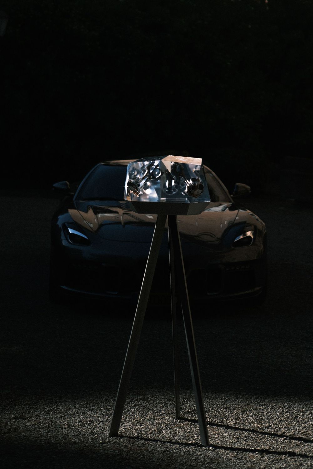 Dark sports car parked at night with a lit display stand in the foreground casting shadows on the ground.