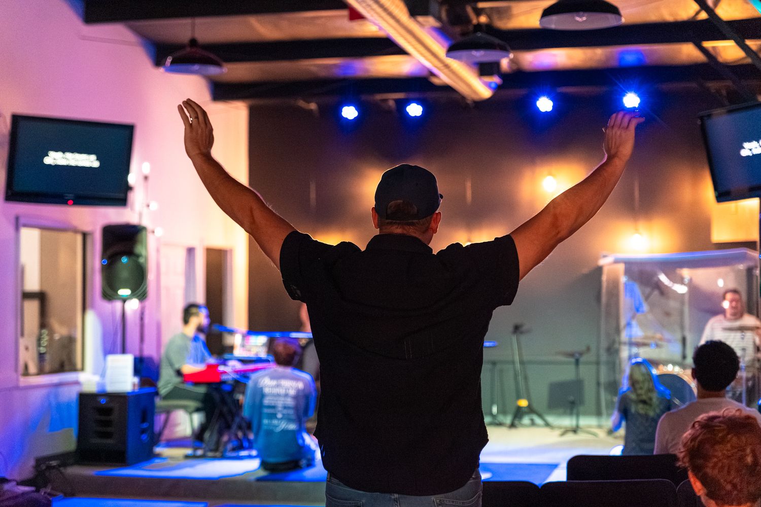Man raises hands in worship at a live church music event with band performing on a colorful lit stage.