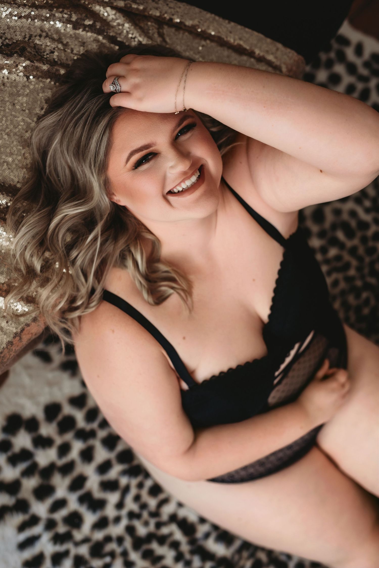 Smiling woman in black lace lingerie lying on leopard print surface with gold sequin backdrop.