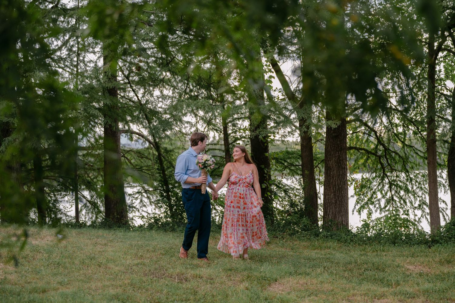 A couple walks hand-in-hand through a lush green park with tall trees and a serene waterfront in the background.