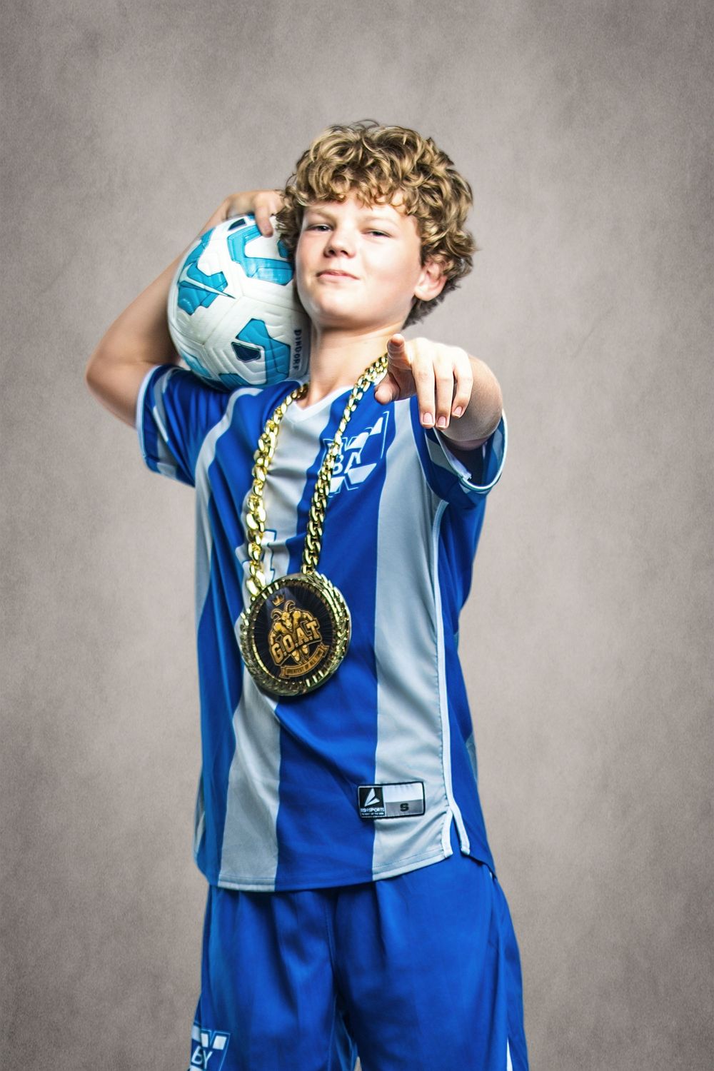 Young boy in blue soccer uniform holding a ball and wearing a gold medal, pointing at camera.