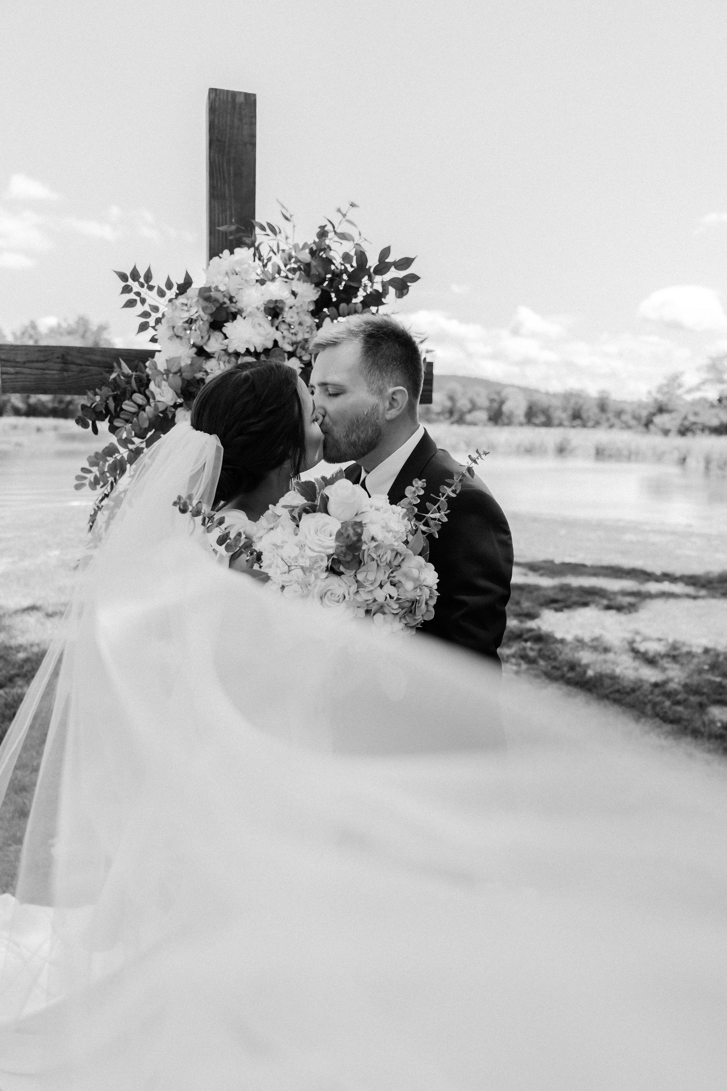 Black and white photo of a bride and groom sharing a kiss at an outdoor wedding ceremony with floral arch and lake backdrop.