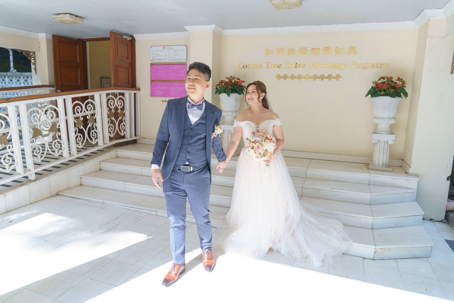 A couple in wedding attire walk down marble stairs in an ornate venue hallway.