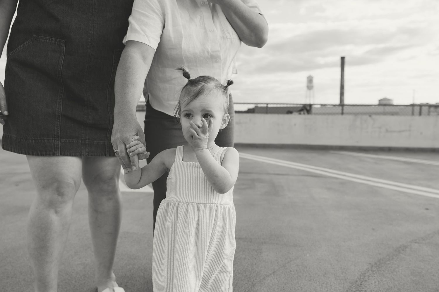 Black and white photo of a toddler girl in white dress holding hands with adult on urban rooftop.