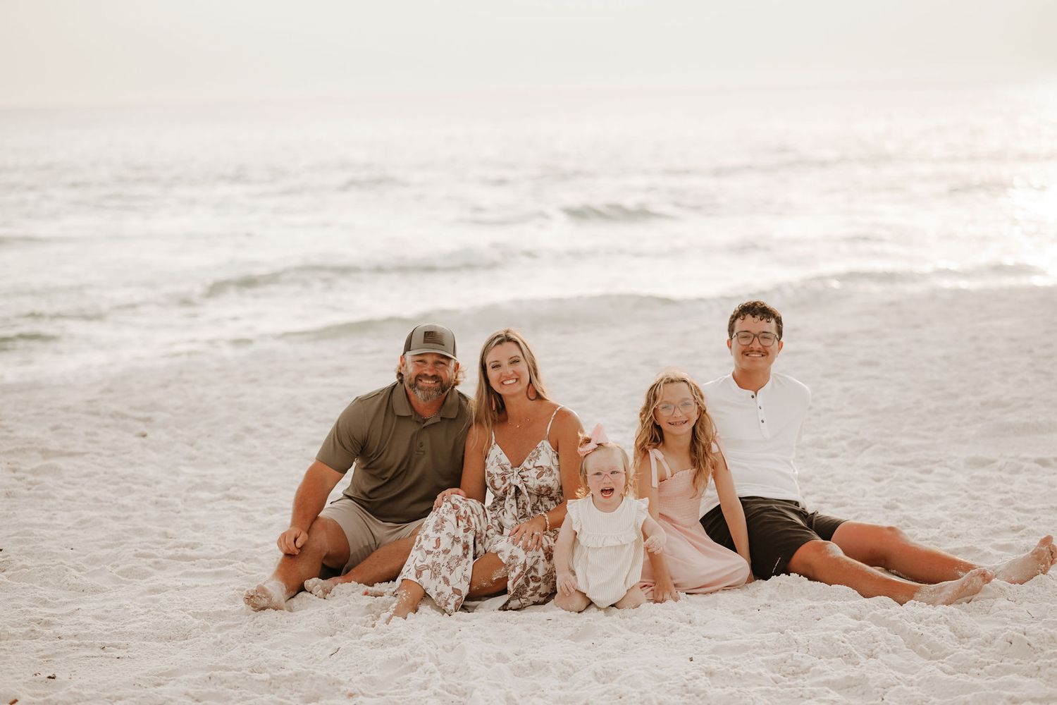 Family of five smiling together on a sandy beach with gentle ocean waves in the background.