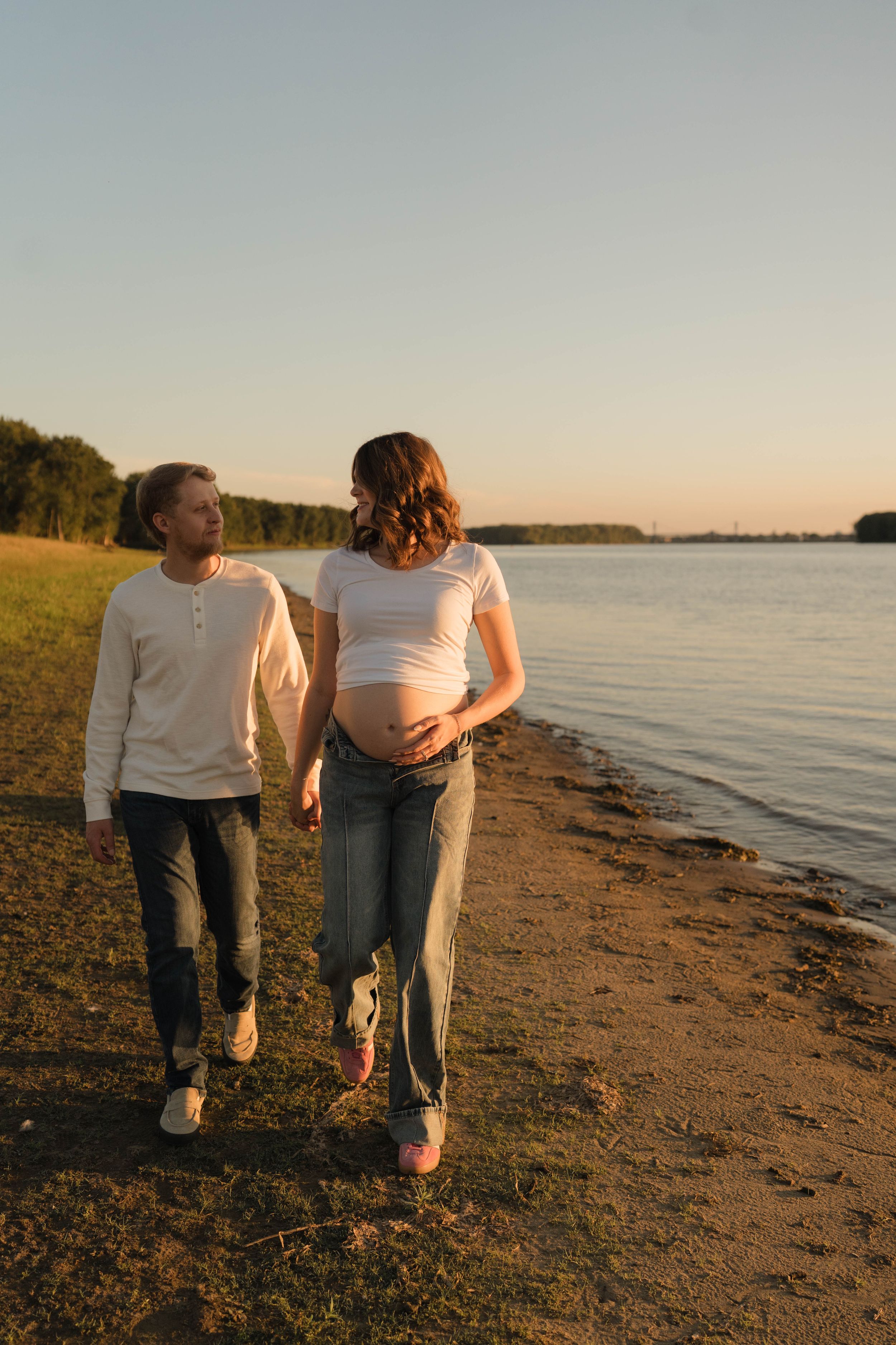 Pregnant couple walking hand in hand along a serene riverside beach at golden sunset hour.