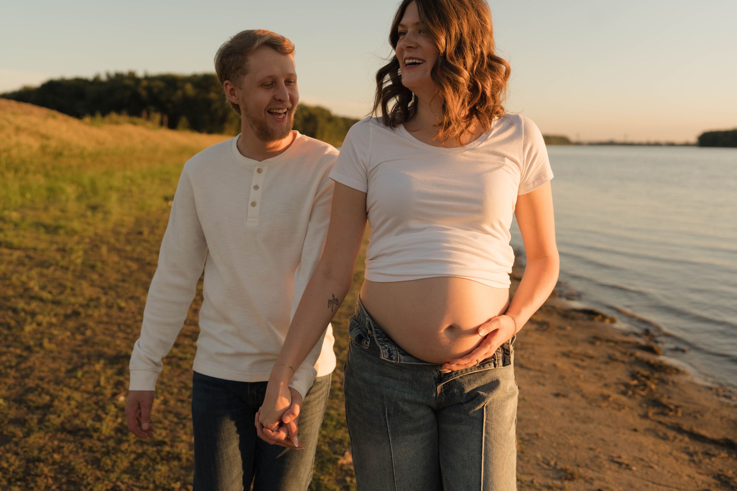 Pregnant woman and man holding hands on a sunny beach at sunset, smiling together in casual white tops and jeans.