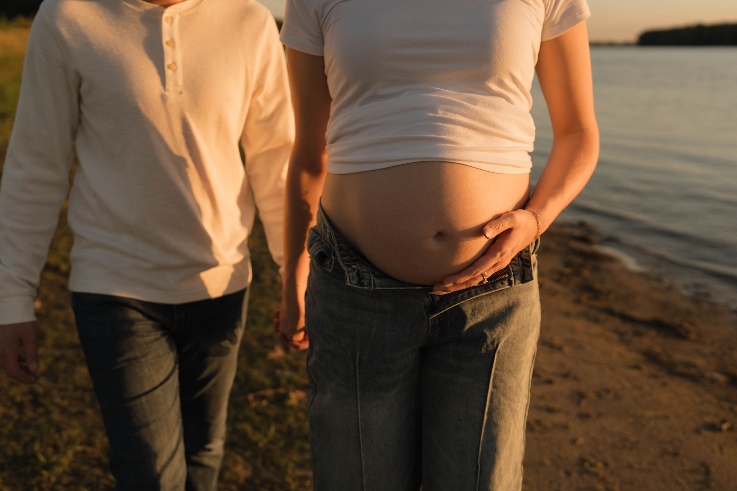 Pregnant woman in white top and jeans showing baby bump near a lake at sunset with partner beside her.