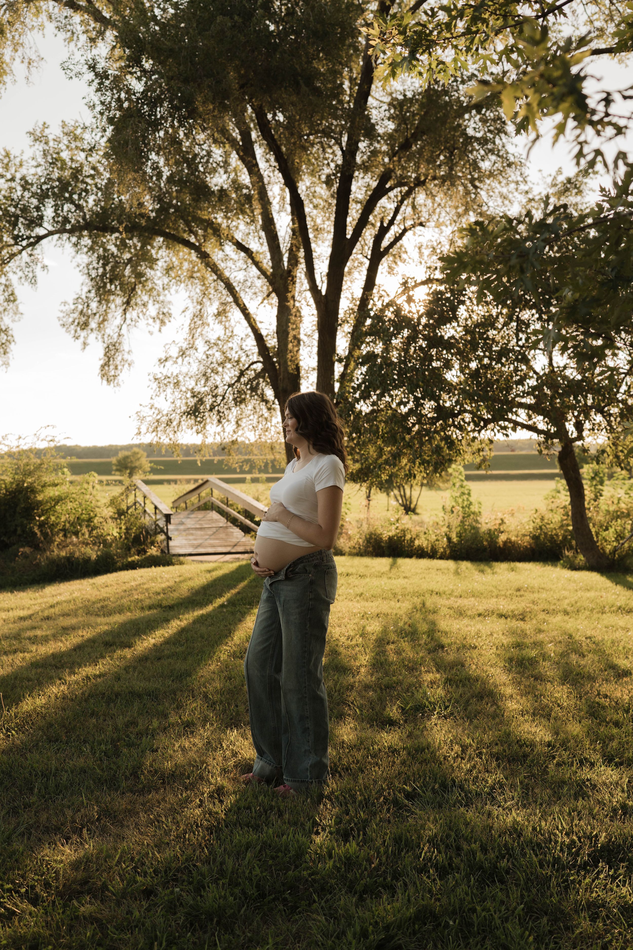 Pregnant woman in white crop top and jeans holding baby bump outdoors at golden hour near large tree.