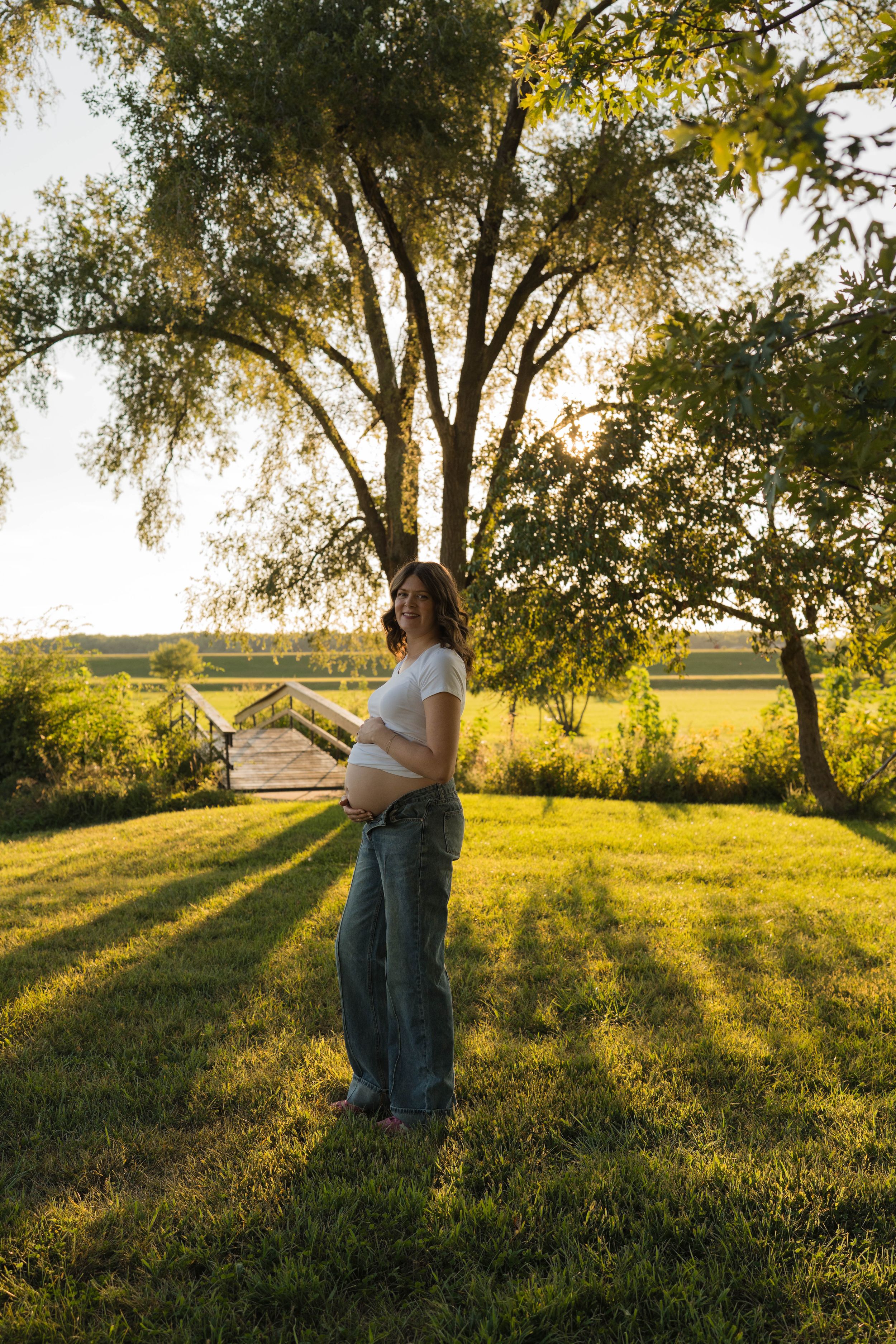 Pregnant woman in white crop top and jeans holding baby bump outdoors in golden hour sunlight near large tree.