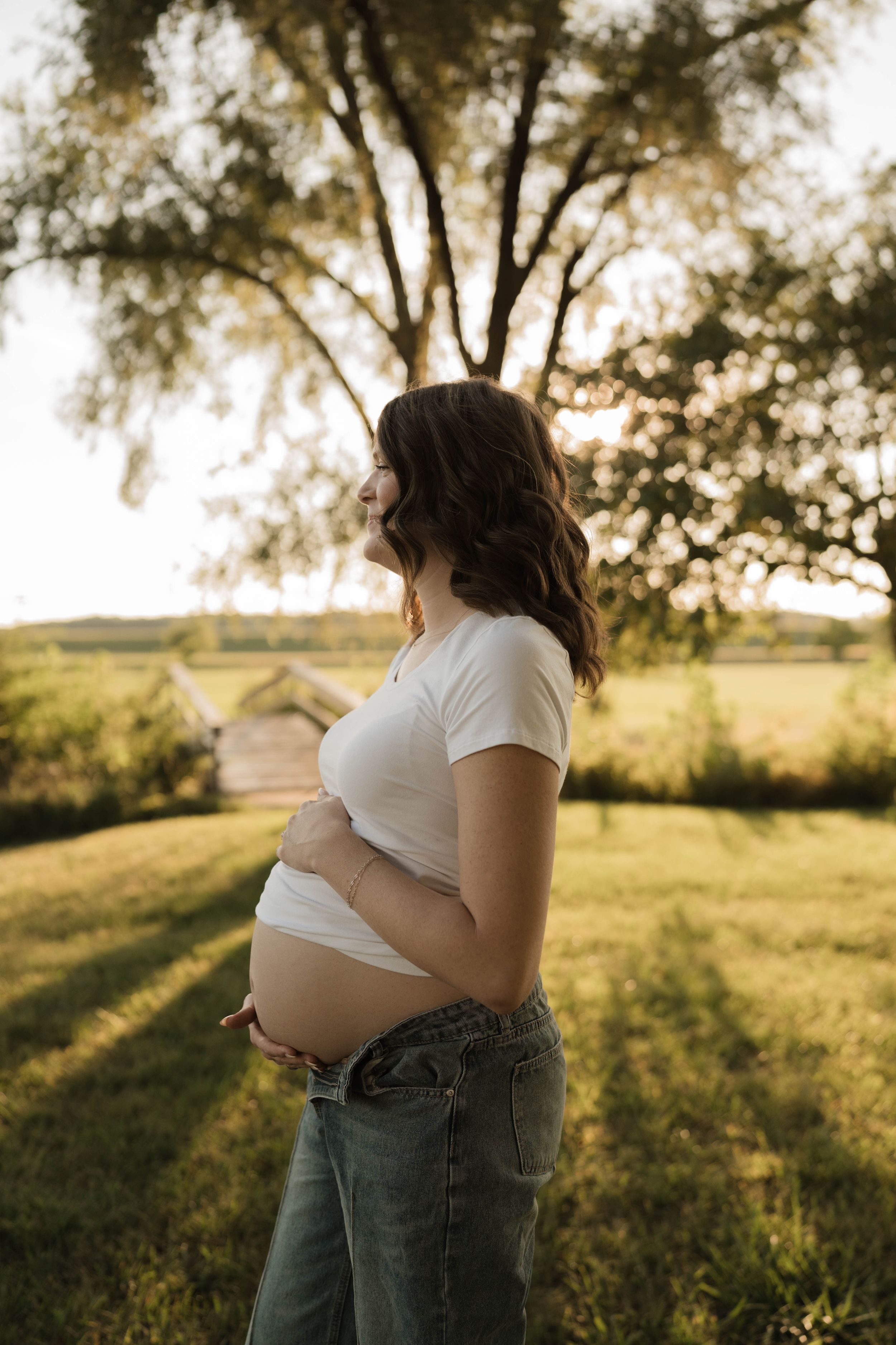 Pregnant woman in white crop top and jeans cradling baby bump outdoors at golden hour.