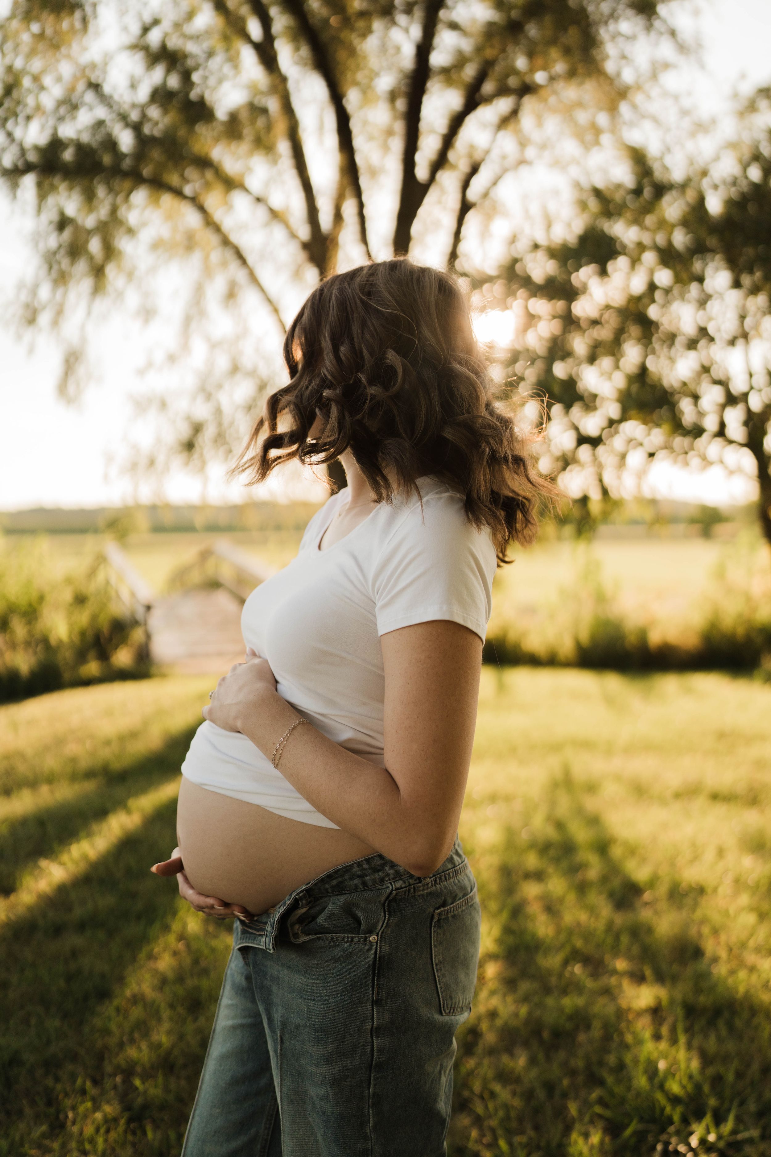 Pregnant woman in white crop top and jeans cradling baby bump outdoors at golden hour near trees.
