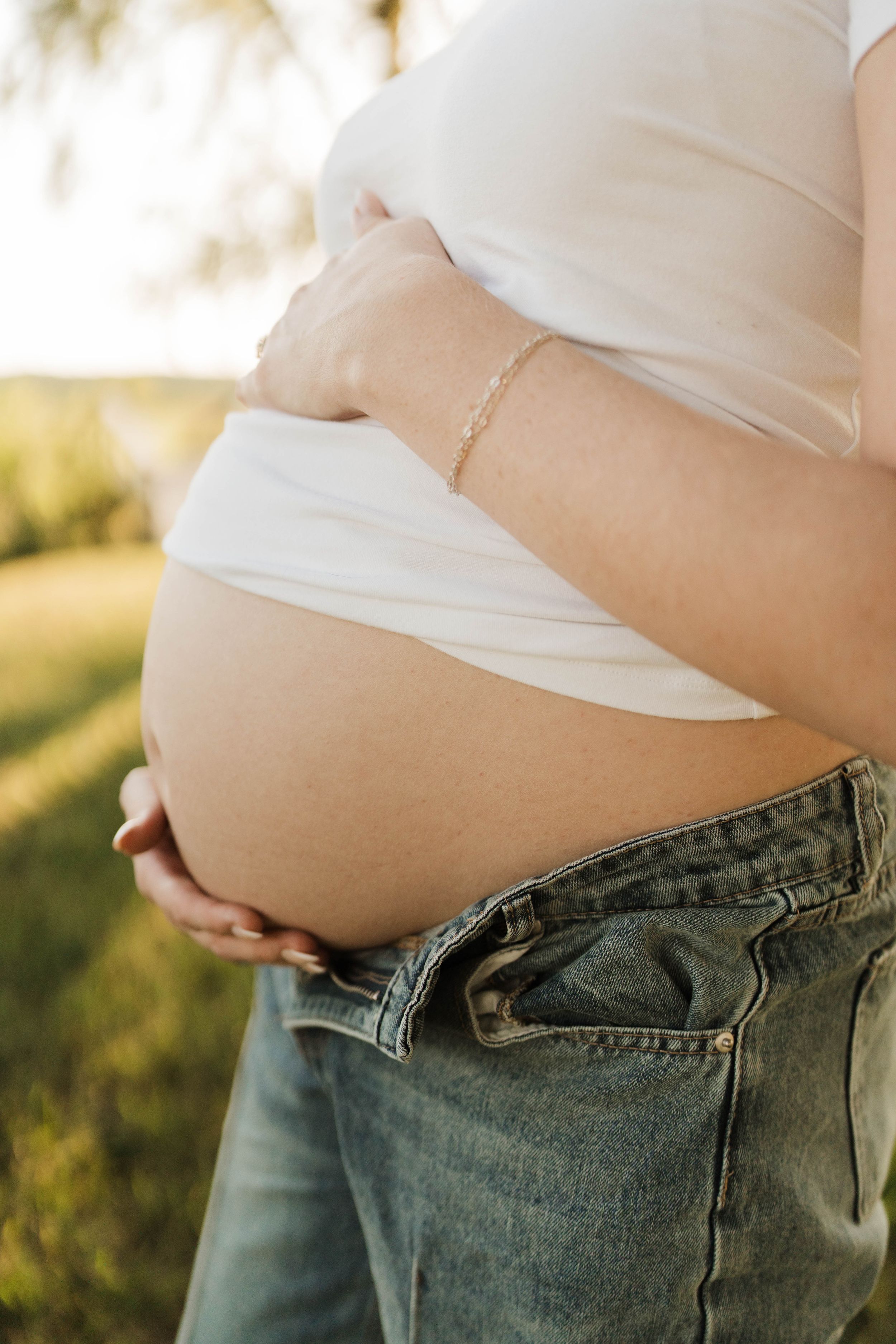 Close-up of a pregnant woman's bare baby bump in jeans and white top, cradled by hands outdoors in golden light.