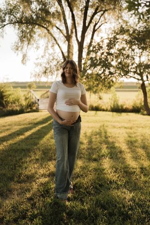 Full length portrait of pregnant woman in white top and jeans standing on green grass at sunset.