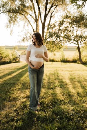 Pregnant woman in white crop top and jeans gently holding belly in golden sunlit meadow.