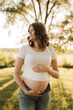 Smiling pregnant woman holding baby bump wearing white tee and jeans in sunny park setting.