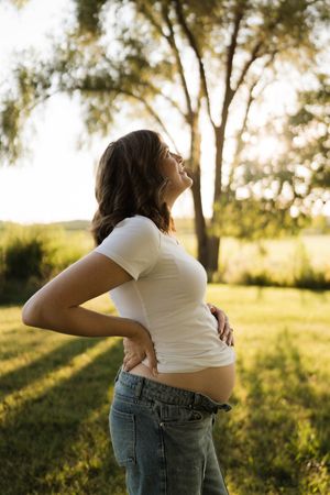 Side profile of pregnant woman arching back holding bump in white top at sunset outdoors.