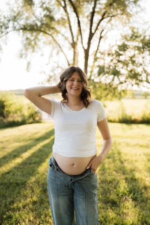 Smiling pregnant woman in white t-shirt and jeans posing outdoors in golden sunlight with green trees behind her.