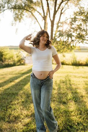 Pregnant woman in white crop top and jeans posing outdoors in golden sunlight with trees in background.