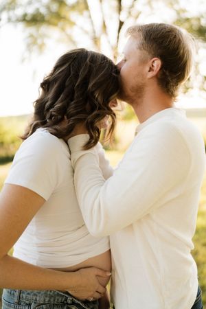 A couple sharing a tender kiss outdoors, both wearing white tops, with a sunlit tree in the background.