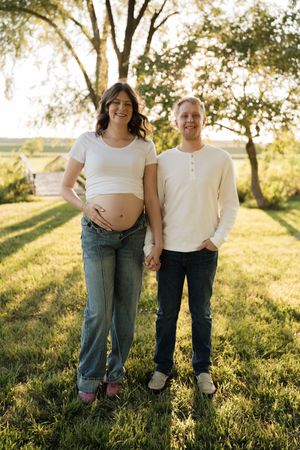 Expecting couple holding hands outdoors at golden hour, woman in white crop top showing baby bump, standing on green grass.