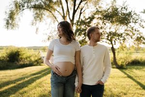 Pregnant couple holding hands outdoors at golden hour, woman cradling baby bump in white crop top and jeans.