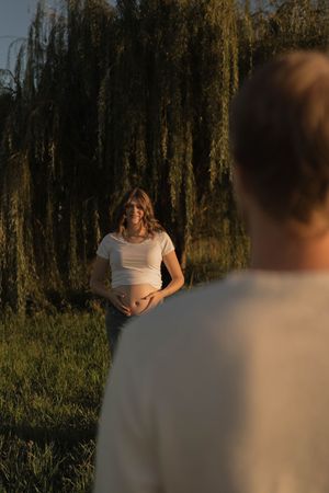 Pregnant woman in white crop top cradling baby bump outdoors near weeping willow tree at golden hour sunset.