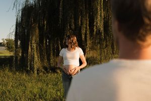 Pregnant woman in white top cradling baby bump outdoors near weeping willow tree in golden sunlight.