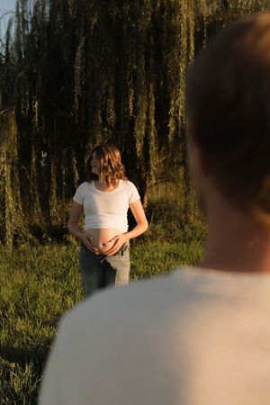 Pregnant woman in white crop top holding baby bump stands near weeping willow tree at sunset, man in foreground.