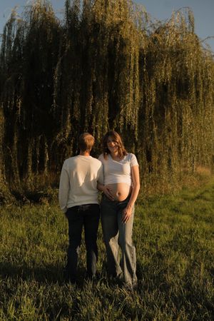Couple standing together near weeping willow tree at sunset, pregnant woman in white top showing baby bump.