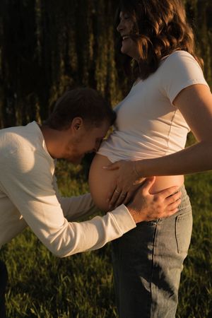 Father-to-be tenderly kissing pregnant partners baby bump outdoors during golden sunset light.