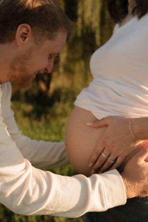 Close-up of man tenderly touching pregnant woman's bare baby bump outdoors near weeping willow in warm sunlight.