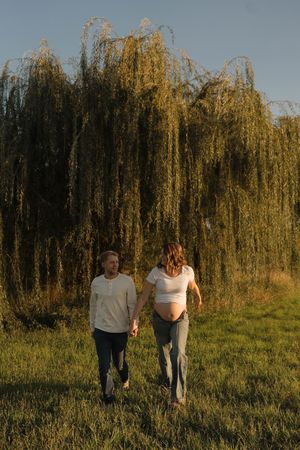 Pregnant couple holding hands walking in a green field in front of a weeping willow tree at golden hour.