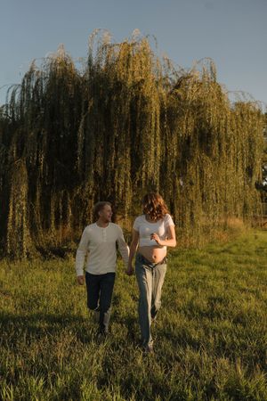 Couple holding hands walking through green field near weeping willow tree during golden sunset, woman visibly pregnant.
